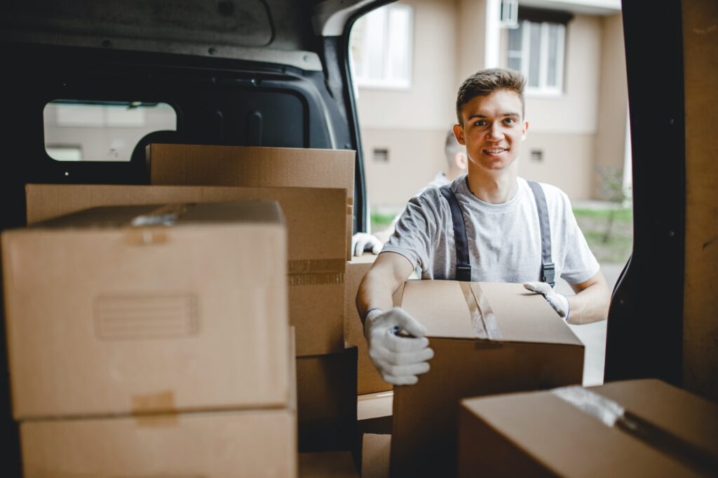 A young handsome smiling worker wearing uniform is standing next to the van full of boxes. House